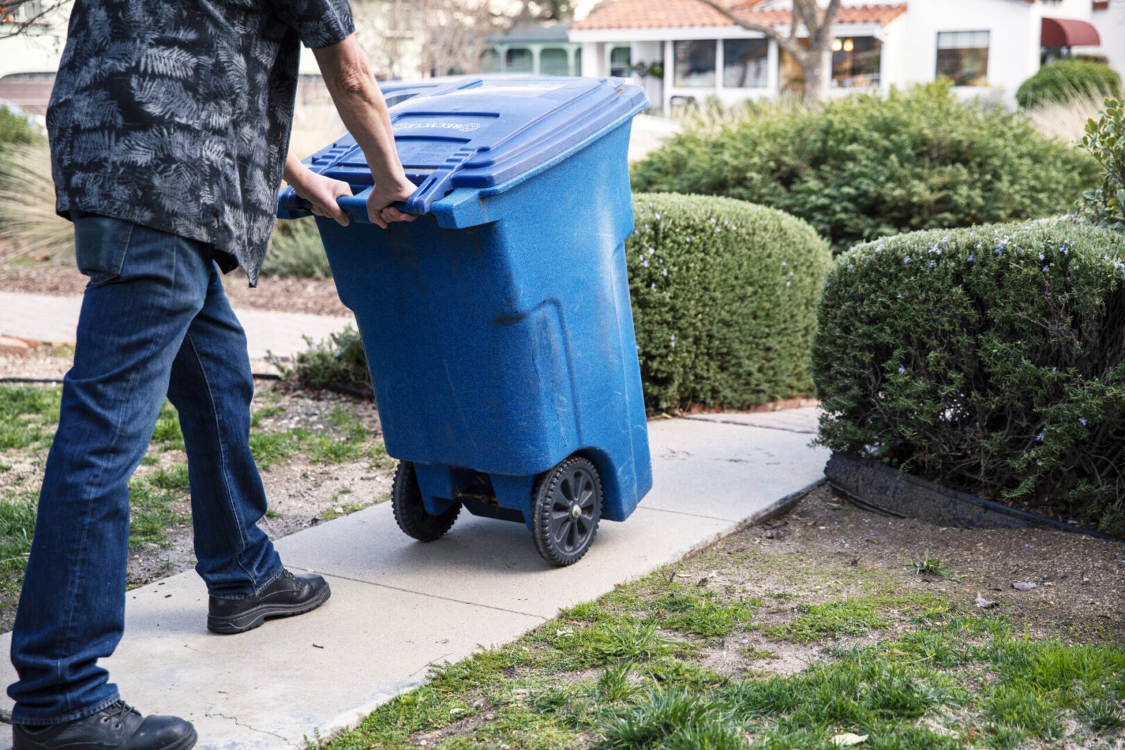 A person pushing a blue trash can down the sidewalk.